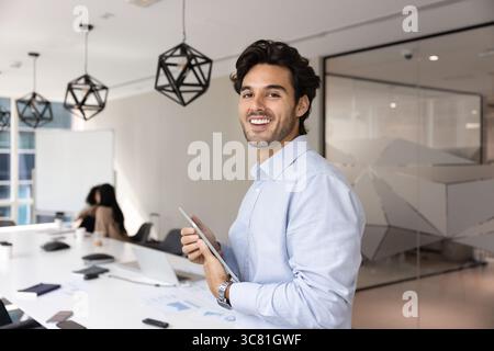 Lächelnder junger männlicher Angestellter, der im Konferenzraum steht und ein Tablet hält Stockfoto