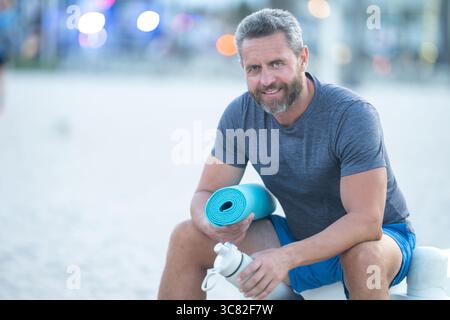 Sport-Fitness-Mann, der draußen auf einer Bank ruht. Reifes Fitnesssportmodell, das nach dem Training auf einer Bank sitzt. Senior Sports man macht danach eine Pause Stockfoto