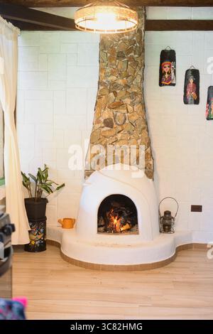 Gemütliches Feuer brennt hell in einem weißen, runden Kamin und schafft eine warme und einladende Atmosphäre in einem rustikalen Wohnzimmer mit Steinkamin und Charme Stockfoto