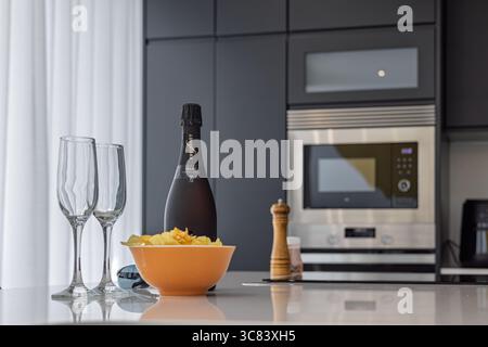 Eine Flasche Prosecco, zwei Gläser und eine Snackschale auf einer glänzend weißen Inseltheke in eleganter schwarzer Küche. Stockfoto