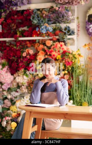 Eine ruhige asiatische Erwachsene Floristin sitzt an einem rustikalen Holztisch in ihrer pulsierenden Blumenboutique inmitten farbenfroher Blüten, die wunderschöne Arrangements kreieren Stockfoto