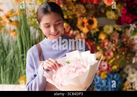 Die lächelnde asiatische Floristin arrangiert sorgfältig schöne rosa Rosen zum Verkauf in einem lebhaften Blumenladen und kreiert wunderschöne frische Sträuße Stockfoto