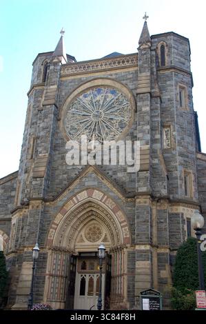 Vorderansicht einer historischen Steinkirche mit einem großen runden Buntglasrosenfenster, einem bogenförmigen Eingang und zwei Türmen unter einem klaren blauen Himmel Stockfoto