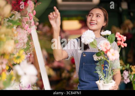 Die lächelnde junge asiatische Floristin arrangiert wunderschöne Blüten in einem lebhaften Einzelhandelsgeschäft. Ihre fröhliche Arbeit schafft angenehme Kundenerlebnisse. Stockfoto