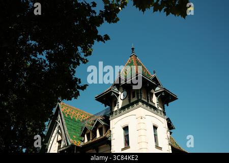 Ein historisches Gebäude in Palic, Serbien, mit einem grün-orange gemusterten Dach und einem kleinen Uhrenturm. Einzigartige architektonische Details unter einem hellen Blau Stockfoto