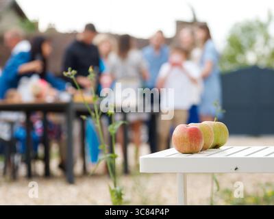 Drei Äpfel ruhen auf einem weißen Gartentisch, während sich im Hintergrund eine verschwommene Familie bei einer goldenen Hochzeitsfeier im versammelt Stockfoto