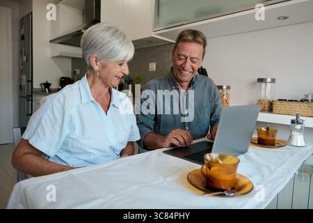 Glückliches Seniorenpaar lacht und benutzt Laptop in der Küche Stockfoto