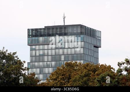 Ein mehrstöckiges modernes Bürogebäude mit einer Fassade aus Glaspaneelen, einer markanten rechteckigen Oberseite und Dachantennen erhebt sich über Baumbewuchs Stockfoto