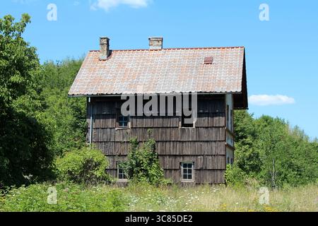Ein altes, zweistöckiges Holzhaus mit einem roten Ziegeldach und zwei Schornsteinen steht zwischen hohem Gras und Wildblumen, umgeben von dichten grünen Bäumen Stockfoto