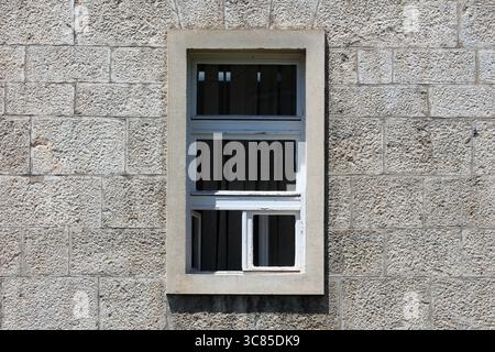 Ein rechteckiges, weiß gerahmtes Fenster mit mehreren geteilten Glasscheiben und einem leicht offenen unteren Paneel ist in eine strukturierte Steinfassade eingefasst Stockfoto