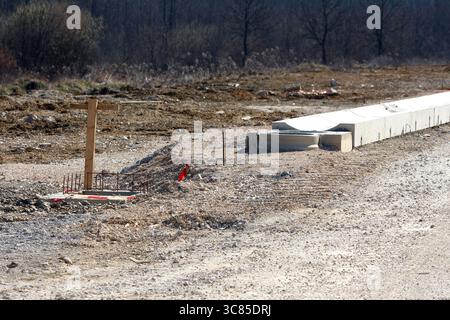 Auf der unbefestigten Straße befindet sich ein hölzerner Übersichtsmarker, der in Beton, freiliegende Bewehrungsstäbe und Betonfertigteile eingebettet ist, um die frühen Phasen der Fahrbahn zu veranschaulichen Stockfoto