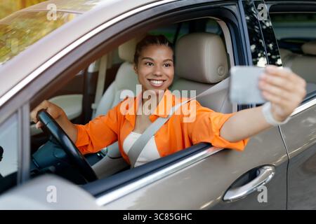 Junge schwarze Frau im Auto lächelt beim Selfie mit Smartphone Stockfoto