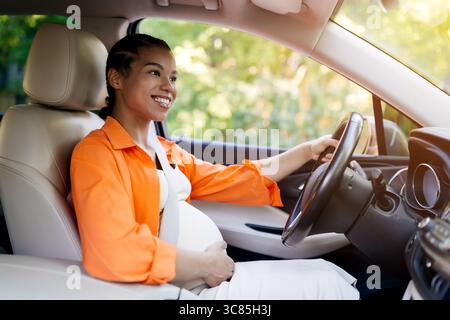 Glückliche schwarze schwangere Frau, die im Sonnenlicht Auto fährt, lächelt und die Fahrt genießt Stockfoto