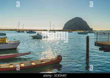 Morro Bay, Kalifornien, USA. Juli 2024. Boote schweben auf glitzerndem Wasser mit Morro Rock im Hintergrund unter hellem Himmel. Stockfoto