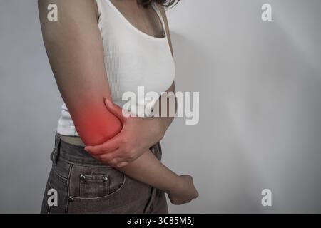 Eine junge Frau in weißem Tanktop und Jeans hält ihren Ellenbogen, der rot hervorgehoben ist, um Schmerzen anzuzeigen. Stockfoto