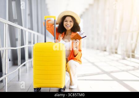 Schöne schwarze Frau kniete neben dem Koffer und hielt Tickets am Flughafen Stockfoto
