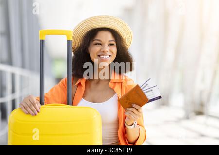 Schwarze Reisende sitzt neben dem Koffer am Flughafen, hält Pass und Tickets Stockfoto