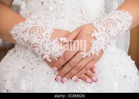 Nahaufnahme der Hände einer Braut, die mit wunderschönen Spitzenhandschuhen und einer stilvollen Maniküre verziert ist. Die weichen Strukturen und die glänzenden Details spiegeln die brida wider Stockfoto