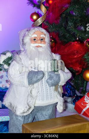Eine fröhliche Weihnachtsmannfigur in Winterkleidung steht neben einem hell erleuchteten Weihnachtsbaum, umgeben von bunten Geschenken und fröhlichen Dekorationen Stockfoto