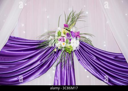 Ein wunderschön gestalteter lila Vorhang mit einer Mischung aus Blumen und Grün schafft eine atemberaubende Kulisse für jede Veranstaltung. Die sanfte Beleuchtung verbessert den Stockfoto