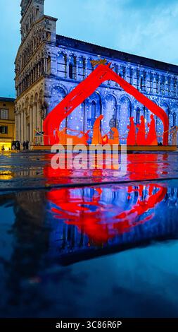 Die beleuchtete Weihnachtskrippe beleuchtet einen Stadtplatz und spiegelt sich lebhaft auf nassem Bürgersteig wider. Die Menschen im Hintergrund bewundern die festliche Ausstellung Stockfoto