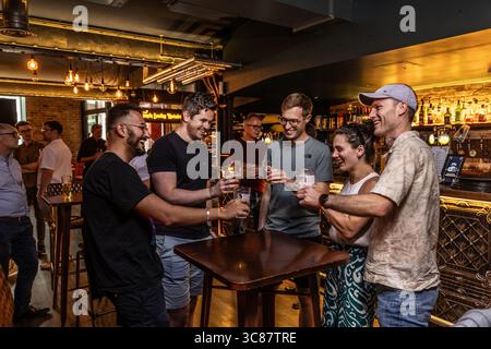 Gruppe junger Profis, die in einer lebhaften Bar Biergläser anklitzen, After-Work-Drinks und Teamarbeit in zwangloser Atmosphäre genießen. Stockfoto