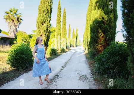 Blonde Frau in blauem Kleid, die im goldenen Sonnenlicht zwischen Zypressen läuft. Stockfoto