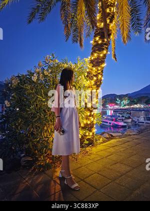 Bezaubernde Abendszene mit einer wunderschönen Frau in einem weißen Kleid, steht an einer Palme mit festlichen Lichtern und Blick auf den lebendigen Moonli Stockfoto