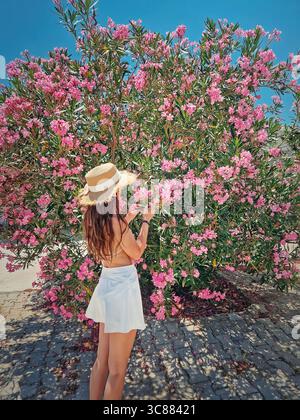 Eine schöne junge Frau mit weißem Rock und Strohhut bewundert einen großen, lebendigen Busch, der mit rosa Nerium Oleander Blumen bedeckt ist Stockfoto