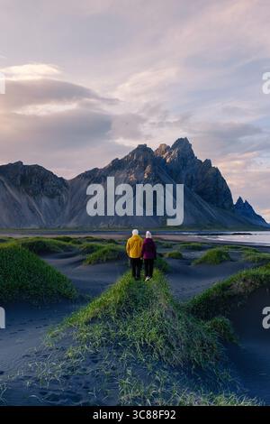 Ein Paar geht Hand in Hand an einem schwarzen Sandstrand Stokksnes Beach Vestrahorn Berg Island, mit beeindruckenden Bergen, die majestätisch im Hintergrund während der goldenen Stunde thronen. Stockfoto