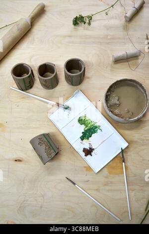 Handbemalte Serviettenringe aus Ton mit botanischen Prägungen im Gange. Pinsel, Farbpalette und Schüssel auf einem Holztisch unterstreichen das künstlerische Stockfoto