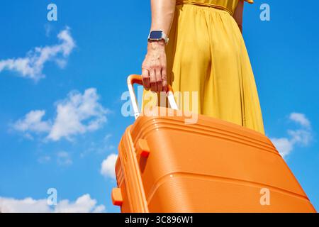 Frau in gelbem Kleid, die einen orangefarbenen Koffer vor blauem Himmel mit Wolken hält. Nahaufnahme einer Reisenden mit Gepäck. Konzept für stilvolles Reisen, va Stockfoto