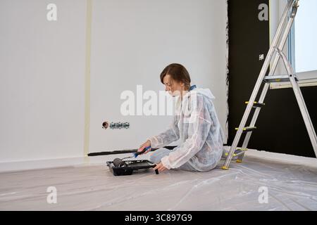 Frau in Schutzanzug Hose weiße Wand mit schwarzem Druck mit Rolle während der Renovierung. Weibliche Arbeiterin, die die Innenwand anstrich. Konzept des Home Impro Stockfoto