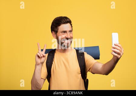 Ein bärtiger Mann macht Selfie mit Rucksack auf gelbem Hintergrund in lässigem Outfit Stockfoto
