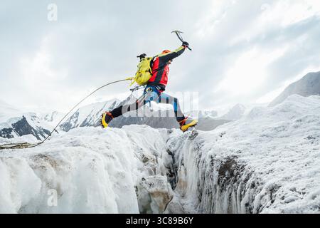 Bergsteiger in pulsierender Ausrüstung hüpft über Gletscherspalten beim Aufstieg des Lenin Gipfels unter klarem Himmel mit schneebedeckten Gipfeln im Hintergrund. Extremer Akt Stockfoto