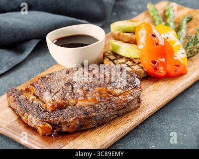 Frisches, heißes Schweinefleisch-Steak auf hölzernem Schneidebrett, serviert Gemüse und Sauce. Köstliches Schweinefleisch auf dunklem Tisch. BBQ Rib Beefsteak oder zartes Schweinefleisch Steak Stockfoto