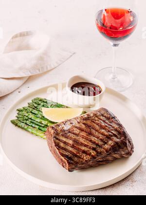 Gegrilltes Fleischsteak auf Teller mit Zitrone und Spargel auf weißem Tischhintergrund. Food and Cuisine Konzept. Verzierung im Restaurant-Stil mit Rotweinglas Stockfoto