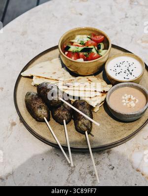 Eine mediterrane Mahlzeit auf einer rustikalen Keramikplatte mit gegrillten Kofta-Spießen, Fladenbrot, einer frischen Gurke und Tomatensalat, Stockfoto