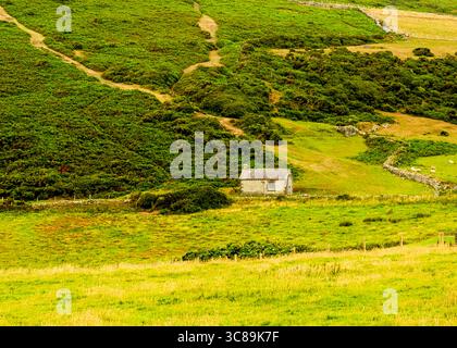 Ein kleines Steingebäude befindet sich auf einem grünen Feld, umgeben von Hügeln, die von Vegetation bedeckt sind. Ein Pfad schlängelt sich den Hügel hinauf. Stockfoto