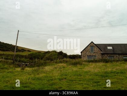 Ein Steinhaus sitzt auf einem Feld aus hohem Gras unter einem bewölkten Himmel. Von der linken Seite des Rahmens erstreckt sich ein Holzmast mit Drähten. Stockfoto