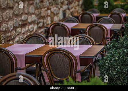 In einem ruhigen Straßencafé in Istanbul, Türkei, stehen Esstische im Freien mit pinken Läufern und gewebten Stühlen an einer rustikalen Steinmauer Stockfoto
