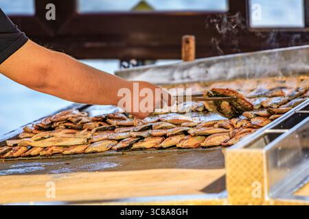 Ein Koch verwendet eine Zange, um frischen Fisch auf einer heißen Grillplatte zu grillen und ihn für ein beliebtes Fisch-Sandwich zu grillen, bei Ofne des Balik Sandwichbootes in Istanbul, Türkei Stockfoto