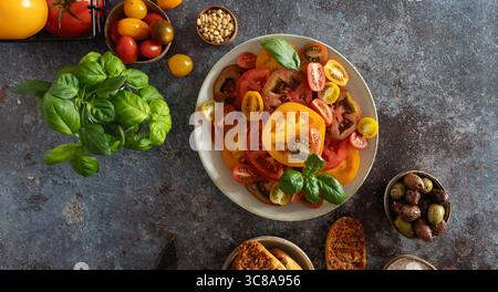 Bunte Tomaten, frische Basilikumstiele, Oliven und Meersalzschale in diagonaler Anordnung auf dunklem rustikalem Hintergrund Stockfoto