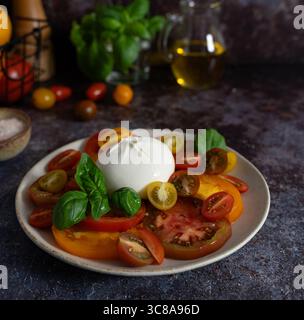 Horizontale Nahaufnahme von gemischten Tomaten mit Burrata in der Mitte, Basilikumblättern, Olivenöl und Schüssel Meersalz auf dunkelblaubraunem Hintergrund Stockfoto