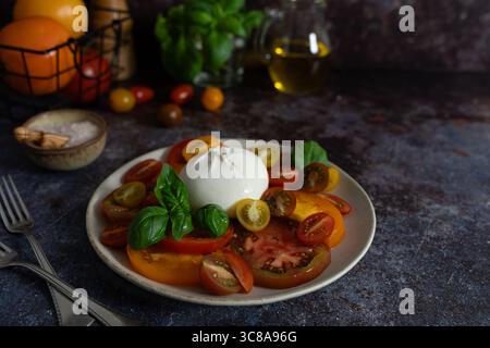 Vorderansicht der Burrata umgeben von bunten Tomaten, frischem Basilikum, Olivenölflasche und Meersalzschale auf rustikalem dunklen Hintergrund Stockfoto