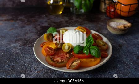 Vorderansicht Nahaufnahme von bunten geschnittenen Tomaten mit Burrata in der Mitte, frischem Basilikum, Olivenölflasche und Schüssel Meersalz auf dunklem Hintergrund Stockfoto
