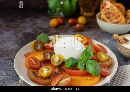 Nahaufnahme von Burrata mit verschiedenen geschnittenen Tomaten, frischem Basilikum und Brot in der Schüssel auf dunkelblaubraunem Hintergrund Stockfoto