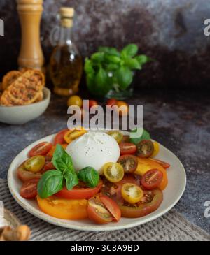 Nahaufnahme von Burrata mit verschiedenen geschnittenen Tomaten, frischem Basilikum, Olivenölflasche und Brot in der Schüssel auf dunkelblaubraunem Hintergrund Stockfoto