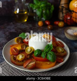Nahaufnahme von Burrata mit geschnittenen und Kirschtomaten, Basilikumblättern, Olivenöl und einer Schüssel Meersalz auf dunkelblaubraunem Hintergrund Stockfoto