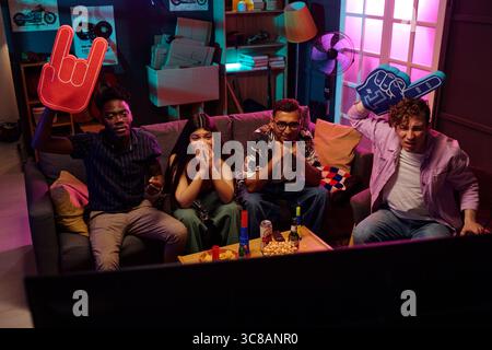 Gruppe junger Erwachsener multiethnischer Freunde, die auf der Couch sitzen und Fernsehen sehen, jubeln und feiern mit Schaumhänden, Snacks und Getränken auf dem Tisch, aufgeregte Gesichtsausdrücke auf den Gesichtern Stockfoto